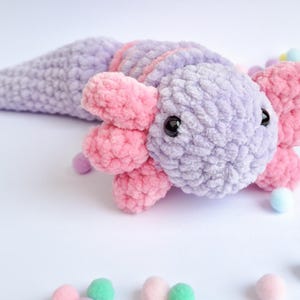 May include: A plush axolotl toy crafted from soft, lavender and pink yarn. The creature features a rounded head with prominent, textured pink external gills on both sides, small black bead eyes, and a long, tapering lavender tail.