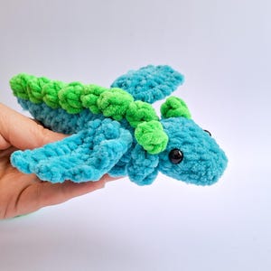 Peut inclure: Un dragon en peluche fait &agrave; la main, turquoise et vert. Le dragon a de grands yeux noirs ronds et de petites ailes. Le dragon est fait d'un fil doux et textur&eacute;. Le dragon est tenu dans une main.