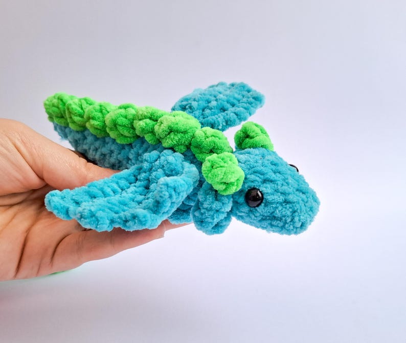 Peut inclure: Un dragon en peluche fait &agrave; la main, turquoise et vert. Le dragon a de grands yeux noirs ronds et de petites ailes. Le dragon est fait d'un fil doux et textur&eacute;. Le dragon est tenu dans une main.