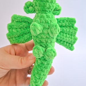 Peut inclure: Un jouet dragon au crochet fait &agrave; la main, vert vif. Le dragon a des ailes textur&eacute;es et duveteuses, ainsi que de petits d&eacute;tails arrondis. Le dragon est tenu dans une main sur un fond blanc. Le corps du dragon est d'une couleur verte vive et unie.