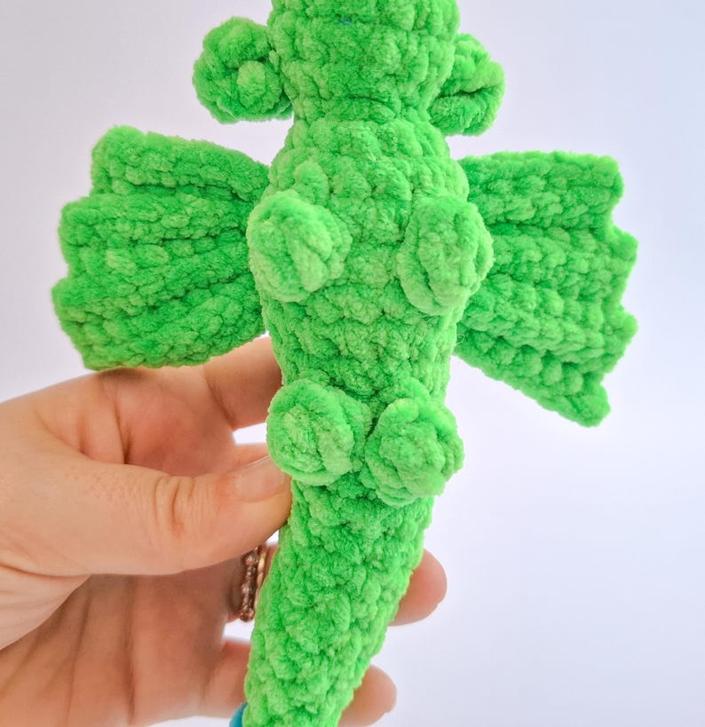 Peut inclure: Un jouet dragon au crochet fait &agrave; la main, vert vif. Le dragon a des ailes textur&eacute;es et duveteuses, ainsi que de petits d&eacute;tails arrondis. Le dragon est tenu dans une main sur un fond blanc. Le corps du dragon est d'une couleur verte vive et unie.