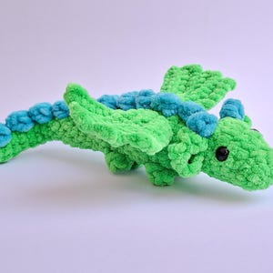 Peut inclure: Un jouet dragon en crochet vert et bleu fait &agrave; la main. Le dragon a une longue queue, des ailes et de petits yeux noirs. Le corps est vert vif, avec des accents bleus sur le dos et la queue.