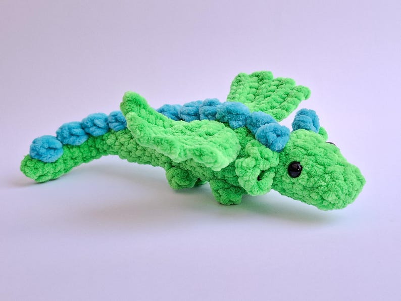 Peut inclure: Un jouet dragon en crochet vert et bleu fait &agrave; la main. Le dragon a une longue queue, des ailes et de petits yeux noirs. Le corps est vert vif, avec des accents bleus sur le dos et la queue.