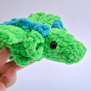 Peut inclure: Un jouet dragon en peluche fait &agrave; la main, vert vif avec des accents bleu sarcelle. Le dragon a un aspect textur&eacute; et flou, un &oelig;il noir et est tenu dans une main. Ce jouet doux convient aux enfants.