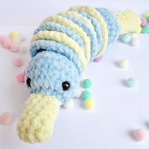 May include: A blue and yellow crocheted plush toy shaped like a platypus. The toy has a long, thin beak and a small, round body. The toy is surrounded by colorful pom-poms.