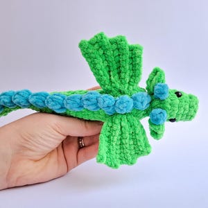 Peut inclure: Un jouet dragon en crochet vert et bleu fait &agrave; la main. Le dragon a un corps vert avec des accents bleus le long de son dos et de sa t&ecirc;te. Le dragon a de petits yeux noirs et des ailes vertes. Le jouet est tenu dans une main.
