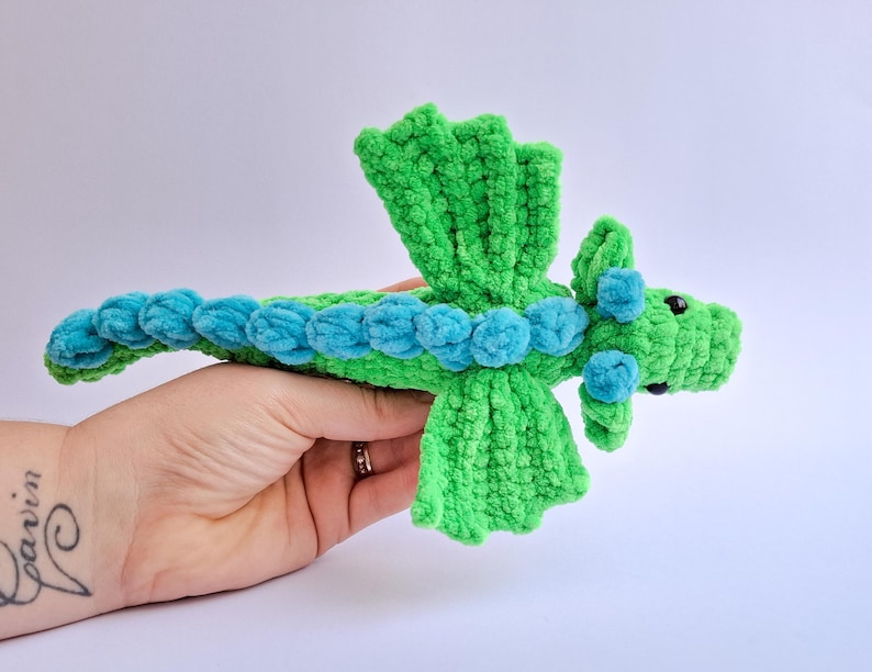 Peut inclure: Un jouet dragon en crochet vert et bleu fait &agrave; la main. Le dragon a un corps vert avec des accents bleus le long de son dos et de sa t&ecirc;te. Le dragon a de petits yeux noirs et des ailes vertes. Le jouet est tenu dans une main.