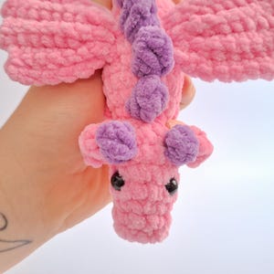 Peut inclure: Un jouet dragon en crochet rose et violet fait &agrave; la main avec des yeux de s&eacute;curit&eacute; noirs. Le dragon a des ailes et des accents violets le long de son dos et de sa t&ecirc;te. La peluche est faite de fil doux et textur&eacute;.