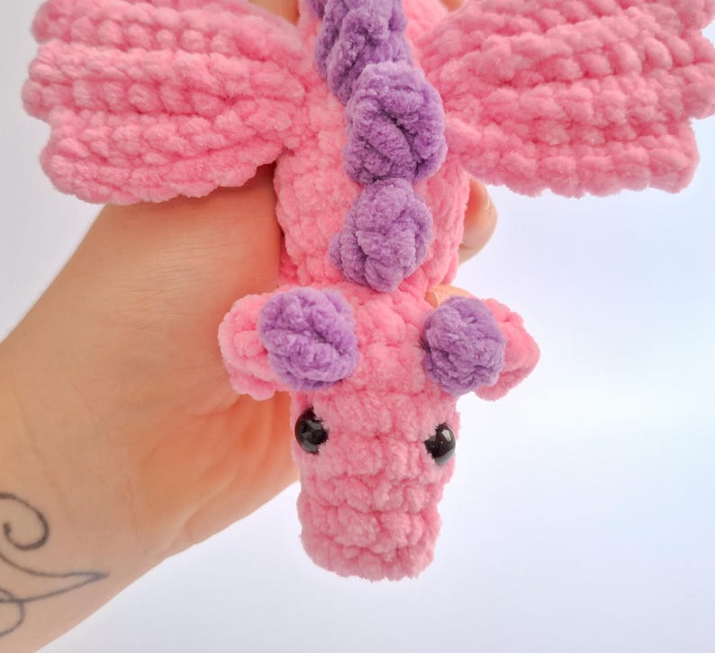 Peut inclure: Un jouet dragon en crochet rose et violet fait &agrave; la main avec des yeux de s&eacute;curit&eacute; noirs. Le dragon a des ailes et des accents violets le long de son dos et de sa t&ecirc;te. La peluche est faite de fil doux et textur&eacute;.
