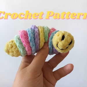 Könnte beinhalten: Ein gehäkelter Regenbogenwurm mit einem gelben Smiley-Gesicht. Der Wurm ist aus weichem, flauschigem Garn gefertigt und etwa 25 cm lang.