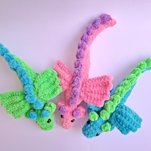 May include: Three handmade crochet dragons in vibrant colors. One is green with blue accents, another is pink with purple, and the third is blue with green. Each dragon has wings and a long, curved neck. These plush toys are perfect for children.