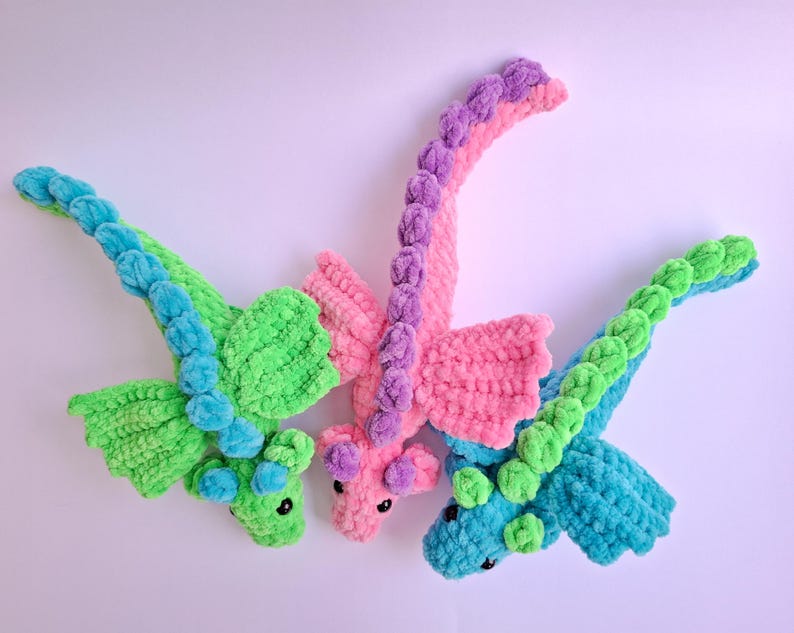 Peut inclure: Trois dragons en crochet faits &agrave; la main aux couleurs vives. L'un est vert avec des d&eacute;tails bleus, un autre est rose avec du violet et le troisi&egrave;me est bleu avec du vert. Chaque dragon a des ailes et un long cou incurv&eacute;. Ces jouets en peluche sont parfaits pour les enfants.