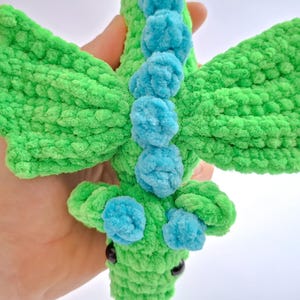 Peut inclure: Un jouet dragon en crochet vert et bleu fait &agrave; la main. Le dragon a de grandes ailes textur&eacute;es et des accents de pompons bleus le long de son dos. Les yeux du dragon sont noirs. Le jouet est fait de fil doux et moelleux.