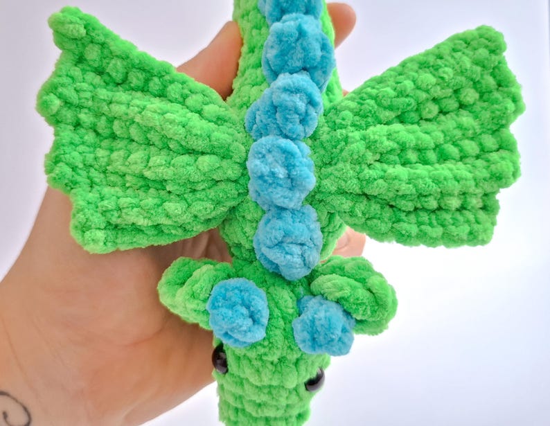 Peut inclure: Un jouet dragon en crochet vert et bleu fait &agrave; la main. Le dragon a de grandes ailes textur&eacute;es et des accents de pompons bleus le long de son dos. Les yeux du dragon sont noirs. Le jouet est fait de fil doux et moelleux.