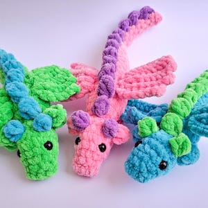 Peut inclure: Trois dragons au crochet faits &agrave; la main aux couleurs vives. L'un est vert et bleu, un autre rose et violet, et le troisi&egrave;me est bleu et vert. Chaque dragon a des ailes et une longue queue incurv&eacute;e. Les dragons sont doux et moelleux.