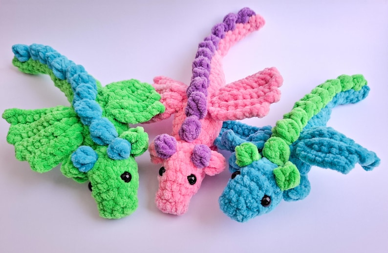 Peut inclure: Trois dragons au crochet faits &agrave; la main aux couleurs vives. L'un est vert et bleu, un autre rose et violet, et le troisi&egrave;me est bleu et vert. Chaque dragon a des ailes et une longue queue incurv&eacute;e. Les dragons sont doux et moelleux.
