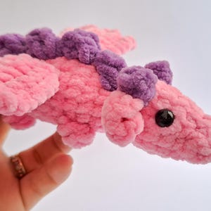 Peut inclure: Un jouet dragon en peluche fait &agrave; la main, rose et violet. Le dragon pr&eacute;sente un corps, des ailes et des &eacute;l&eacute;ments d&eacute;coratifs textur&eacute;s et flous. Il a un &oelig;il noir et est con&ccedil;u pour &ecirc;tre doux et c&acirc;lin.