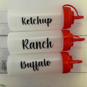 May include: Three white plastic squeeze bottles with red caps, labeled "Ketchup", "Ranch", and "Buffalo" in black script. The bottles are designed for dispensing condiments.