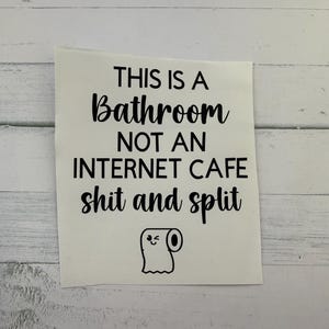 May include: A white sticker with black text that reads "THIS IS A BATHROOM NOT AN INTERNET CAFE shit and split." A cartoon toilet paper roll with a winking face is at the bottom.
