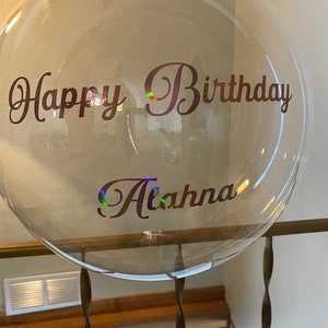 May include: A clear balloon with the text "Happy Birthday Alahna" written in brown cursive lettering.