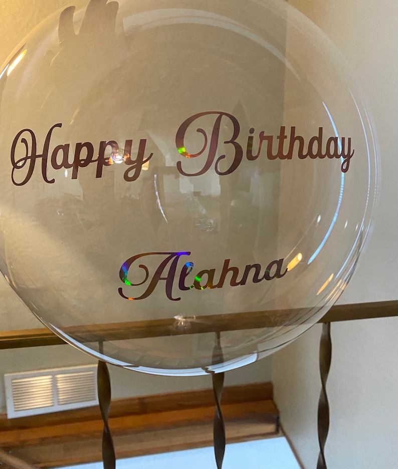 May include: A clear balloon with the text "Happy Birthday Alahna" written in brown cursive lettering.