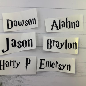 May include: Six white rectangular stickers with black text. The stickers display the names "Dawson", "Alahna", "Jason", "Braylon", "Harry P", and "Emersyn" in a stylized font. The stickers are arranged on a light-colored wooden surface.
