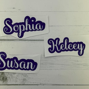 May include: Three custom name decals in a dark purple script font. The names are "Sophia", "Kelsey", and "Susan", each on a white background. The decals are displayed on a light wood-grain surface.
