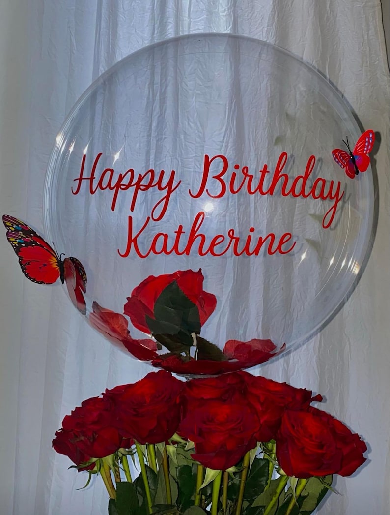 May include: A clear balloon with the text "Happy Birthday Katherine" in red lettering. Two red butterflies are attached to the balloon. The balloon is filled with red rose petals and sits on top of a bouquet of red roses.