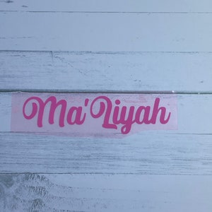 May include: Pink vinyl lettering on a white wood background that reads "Ma'Liyah".
