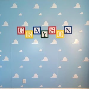 May include: A light blue wall with white cloud stickers and the name "GRAYSON" spelled out in colorful block letters.