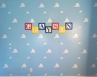 Puede incluir: Una pared azul claro con pegatinas de nubes blancas y el nombre "GRAYSON" escrito en letras de bloques de colores.