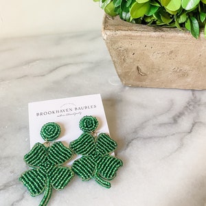 May include: Green beaded clover earrings with gold accents. The earrings are on a white card with the text "BROOKHAVEN BAUBLES" and "handmade statement jewelry".