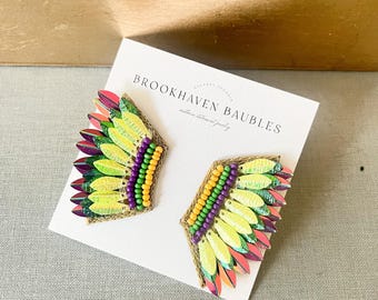 May include: A pair of colorful beaded earrings with a feather design. The earrings are made with green, yellow, purple, and pink beads. The earrings are displayed on a white card with the text "BROOKHAVEN BAUBLES" printed on it.