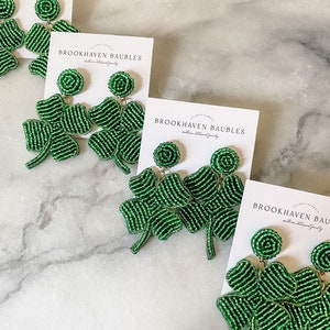 May include: Four pairs of green beaded shamrock earrings with gold accents. The earrings are displayed on white marble surfaces with cards that read "Brookhaven Baubles".