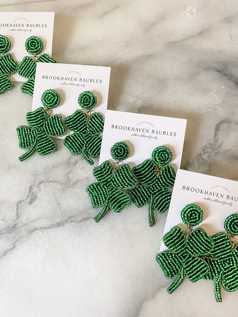 May include: Four pairs of green beaded shamrock earrings with gold accents. The earrings are displayed on white marble surfaces with cards that read "Brookhaven Baubles".