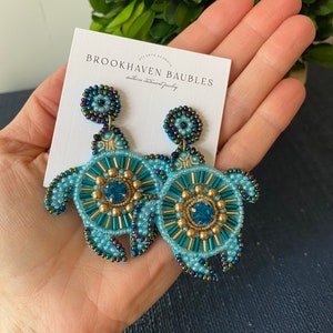 May include: A pair of turquoise and gold beaded earrings shaped like sea turtles. The earrings feature a large turquoise gemstone in the center of each turtle. The earrings are handmade and have a bohemian style. The earrings are from the shop Brookhaven Baubles.