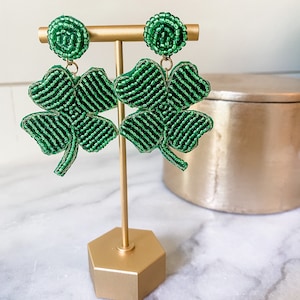 May include: A pair of green beaded shamrock earrings on a gold stand. The earrings are made of small green beads and are shaped like four-leaf clovers.