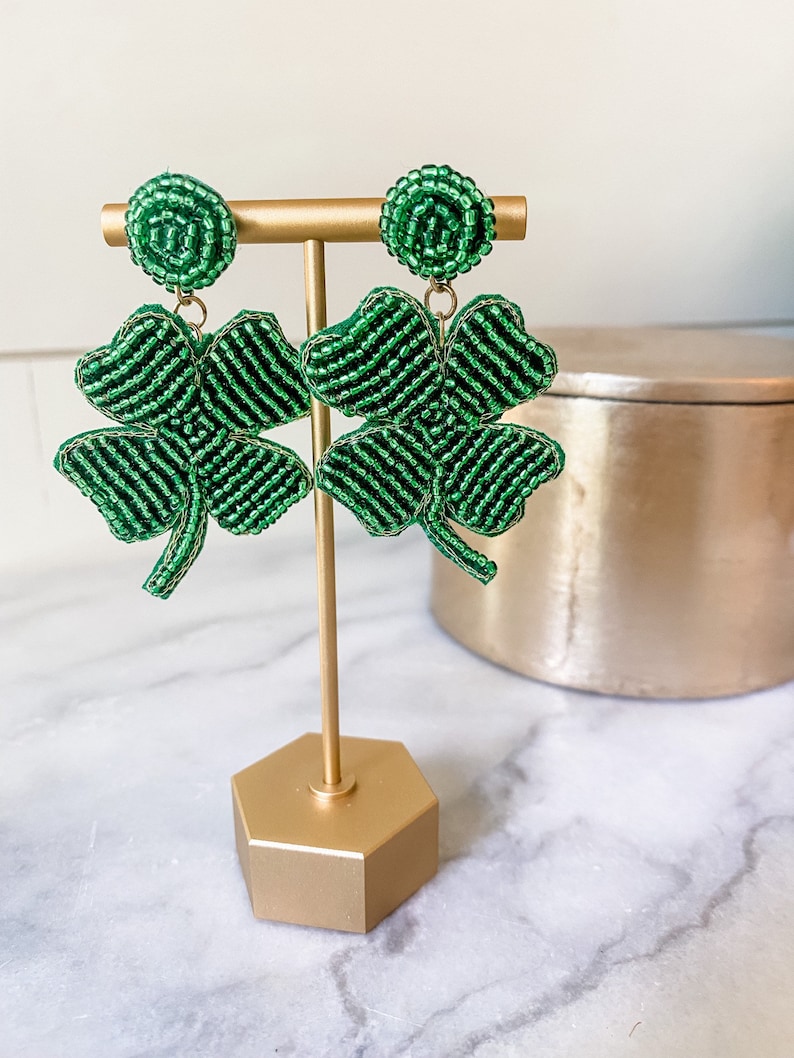 May include: A pair of green beaded shamrock earrings on a gold stand. The earrings are made of small green beads and are shaped like four-leaf clovers.