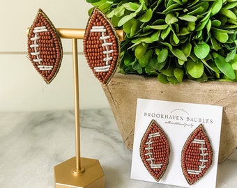 May include: A pair of brown and white beaded football earrings. The earrings are displayed on a gold stand and a white card with the text "Brookhaven Baubles".