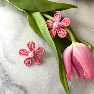 May include: Two pink flower-shaped earrings with a pearl center, displayed on a white surface with a pink tulip and green leaves.