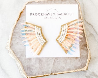 May include: A pair of gold and iridescent pink feather earrings with white pearl accents. The earrings are displayed on a white card with the text "BROOKHAVEN BAUBLES southern statement jewelry".