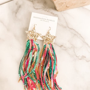May include: Gold star-shaped earrings with long, colorful tassels made of sequins. The tassels are a mix of pink, green, and gold sequins. The earrings are labeled "Brookhaven Baubles".