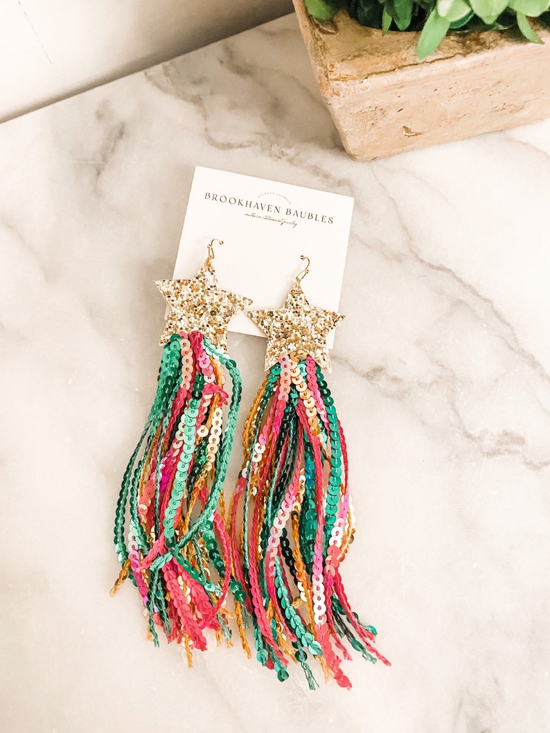 May include: Gold star-shaped earrings with long, colorful tassels made of sequins. The tassels are a mix of pink, green, and gold sequins. The earrings are labeled "Brookhaven Baubles".