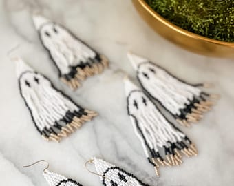 May include: Four pairs of beaded ghost earrings. The earrings are white with black details and gold accents. The ghosts have a simple, smiling face.