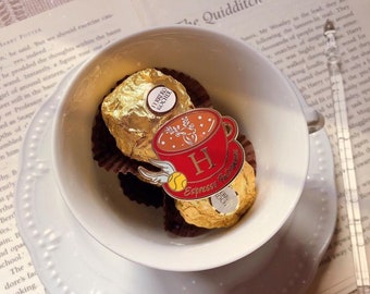 May include: A white ceramic teacup and saucer with a red and gold enamel pin featuring a coffee cup with a letter H and the text "Espresso Patronum". The pin is sitting on top of two chocolate candies wrapped in gold foil.