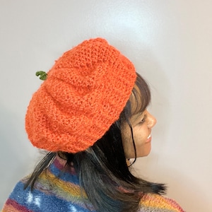 May include: A crocheted pumpkin hat in orange with a green stem. The hat is being worn by a person with dark hair.