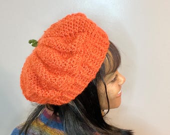 May include: A crocheted pumpkin hat in orange with a green stem. The hat is being worn by a person with dark hair.