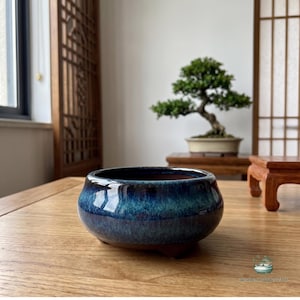 Può includere: Un vaso per bonsai rotondo in ceramica con una smaltatura lucida che sfuma dal blu marino profondo al turchese vivace. Il vaso presenta una silhouette liscia e curva con un'ampia apertura e piccoli piedini alla base, con una finitura riflettente.