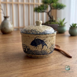 May include: A round ceramic lidded jar with a speckled beige finish, featuring dark blue hand-painted illustrations of a fish and stylized waves. The jar has a matching lid with a small knob handle and is accompanied by a small wooden spoon.