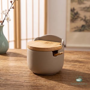 May include: A matte light gray ceramic container with a circular bamboo lid and a small wooden spoon tucked into a side notch. The lid features a small engraved square pattern. The container has a smooth, rounded base and a slightly raised back rim.